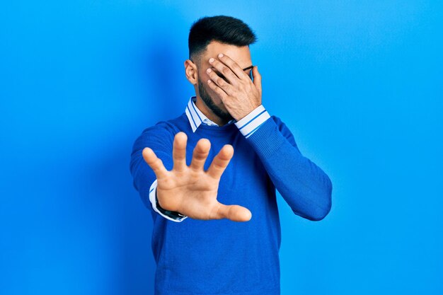 Jeune homme hispanique avec barbe portant un chandail bleu décontracté couvrant les yeux avec les mains et faisant un geste d'arrêt avec une expression de tristesse et de peur. concept embarrassé et négatif.
