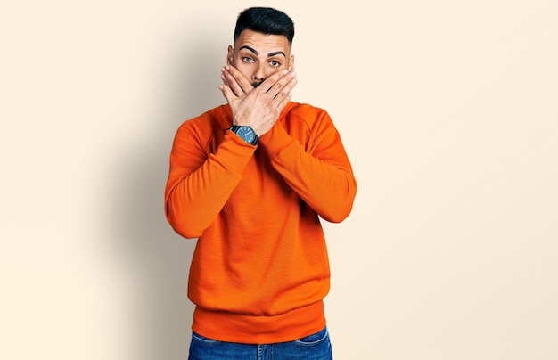 Jeune homme hispanique avec barbe portant un chandail orange décontracté choqué couvrant la bouche avec les mains par erreur. notion secrète.