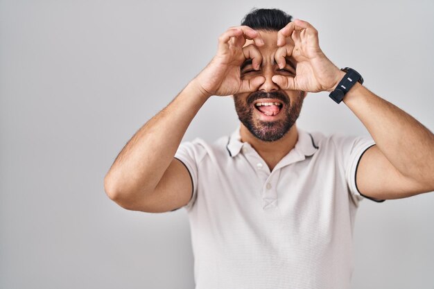 Jeune homme hispanique avec barbe portant des vêtements décontractés sur fond blanc faisant un geste correct comme des jumelles tirant la langue des yeux regardant à travers les doigts expression folle