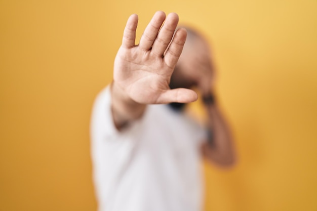 Photo gratuite jeune homme hispanique avec barbe et tatouages debout sur fond jaune couvrant les yeux avec les mains et faisant un geste d'arrêt avec une expression triste et de peur concept embarrassé et négatif