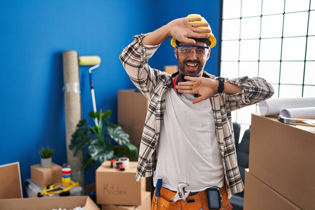 Jeune homme hispanique avec barbe travaillant à la rénovation de la maison souriant joyeux jouant peek a boo avec les mains montrant le visage surpris et sorti