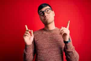 Photo gratuite jeune homme hispanique beau portant des lunettes nerd sur fond rouge pointant vers le haut l'air triste et contrarié indiquant la direction avec les doigts mécontents et déprimés