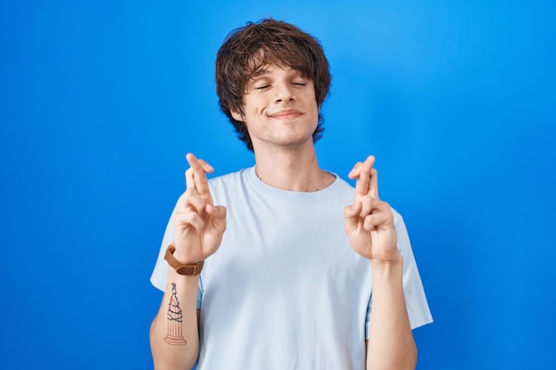 Photo gratuite jeune homme hispanique debout sur fond bleu gesticulant doigt croisé souriant avec espoir et les yeux fermés chance et concept superstitieux