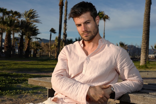 Jeune homme hispanique portant une chemise rose et posant sur la plage près des palmiers