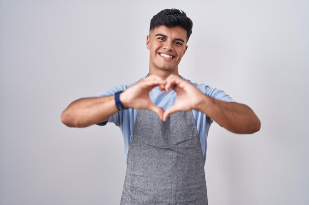 Jeune homme hispanique portant un tablier sur fond blanc souriant amoureux faisant la forme de symbole de coeur avec les mains. notion romantique.