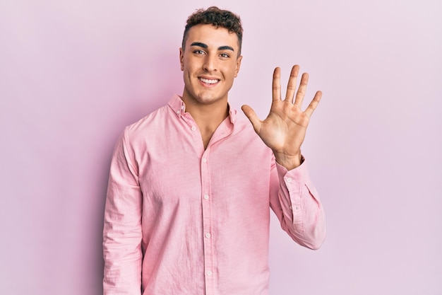 Jeune homme hispanique portant des vêtements décontractés montrant et pointant vers le haut avec les doigts numéro cinq tout en souriant confiant et heureux