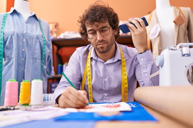 Jeune homme hispanique tailleur écoutant un message vocal par smartphone dessinant la conception de vêtements à l'atelier