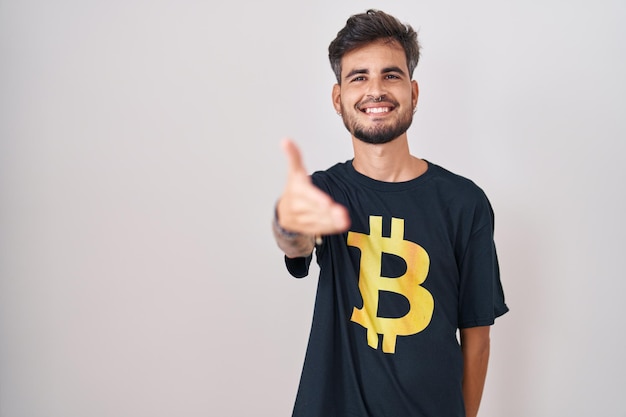 Photo gratuite jeune homme hispanique avec des tatouages portant un t-shirt bitcoin souriant amical offrant une poignée de main comme salutation et accueillant une entreprise prospère