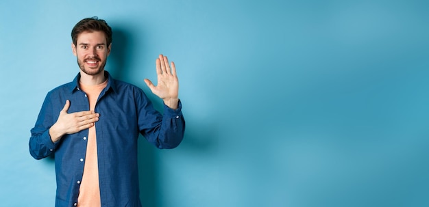 Photo gratuite un jeune homme honnête souriant et faisant la promesse tenant la main sur le cœur et le bras levé s'engage ou prête serment