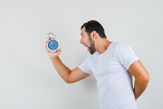 Jeune homme hurlant en regardant l'horloge en t-shirt blanc et à la panique, vue de face.