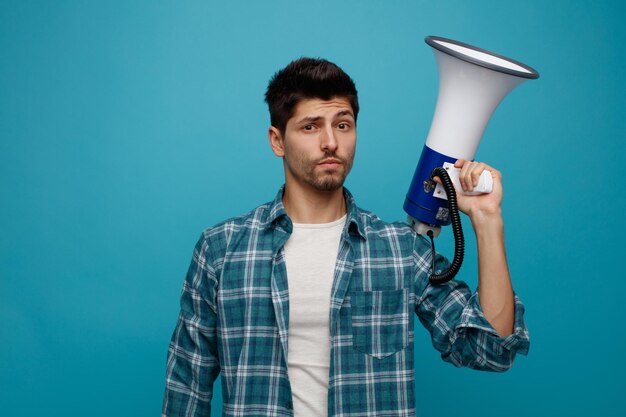 Jeune homme incertain regardant la caméra tenant le haut-parleur isolé sur fond bleu