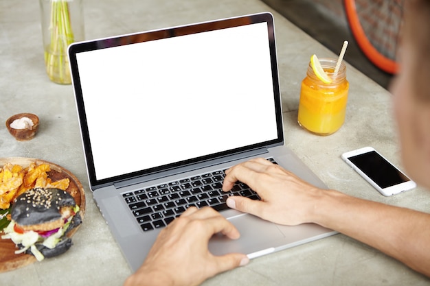 Jeune homme indépendant travaillant sur son projet sur un ordinateur portable générique assis au café et utilisant le Wi-Fi gratuit. Étudiant de sexe masculin naviguant sur Internet ou vérifiant le courrier électronique sur un appareil électronique pendant le déjeuner
