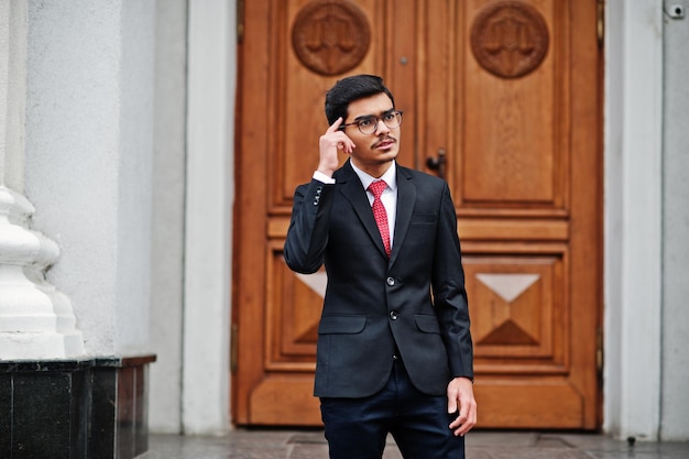 Jeune homme indien portant des lunettes sur un costume noir avec une cravate rouge posée en plein air contre la porte du bâtiment montre que sa pensée