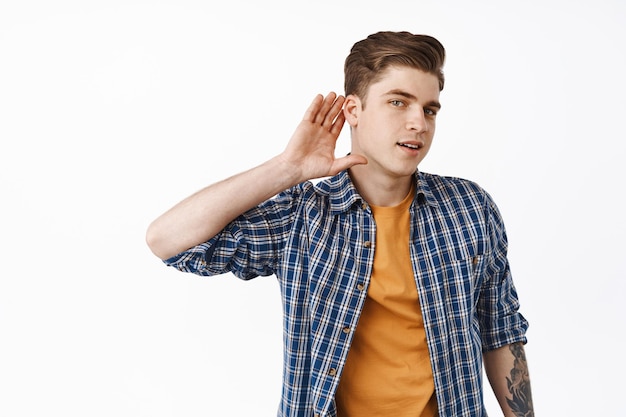 Un jeune homme intrigué écoutant, un garçon entend des commérages, tient la main près de l'oreille et essaie d'entendre la conversation, debout sur fond blanc