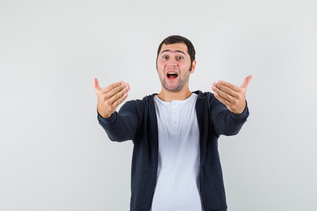 Jeune homme invitant à venir en t-shirt blanc et sweat à capuche noir à glissière sur le devant et à l'optimiste, vue de face.