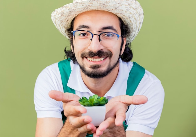Photo gratuite jeune homme de jardinier barbu portant combinaison et chapeau montrant plante en pot à l'avant souriant joyeusement debout sur un mur vert clair