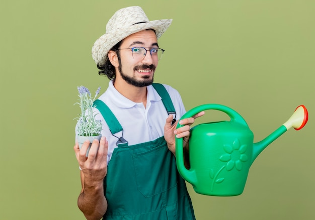 Jeune homme de jardinier barbu portant une combinaison et un chapeau tenant un arrosoir et une plante en pot à l'avant souriant avec un visage heureux debout sur un mur vert clair