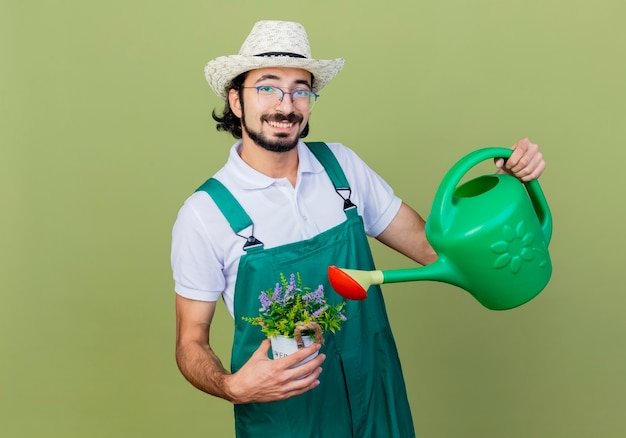 Jeune homme de jardinier barbu portant une combinaison et un chapeau tenant un arrosoir et une plante en pot à l'avant souriant avec un visage heureux debout sur un mur vert clair