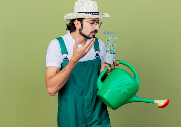 Jeune homme de jardinier barbu portant une combinaison et un chapeau tenant un arrosoir et une plante en pot sentant une odeur agréable debout sur un mur vert clair