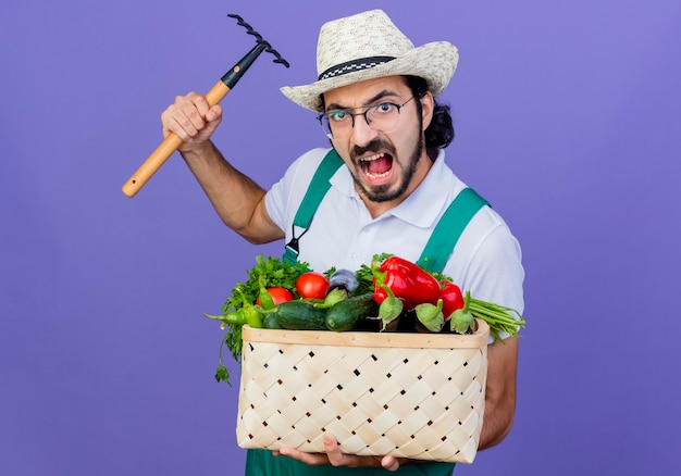 Photo gratuite jeune homme de jardinier barbu portant combinaison et chapeau tenant caisse pleine de légumes balançant un mini râteau avec visage en colère debout sur le mur bleu