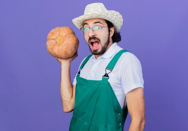 Jeune homme de jardinier barbu portant combinaison et chapeau tenant la citrouille à l'avant en criant d'être excité debout sur le mur bleu