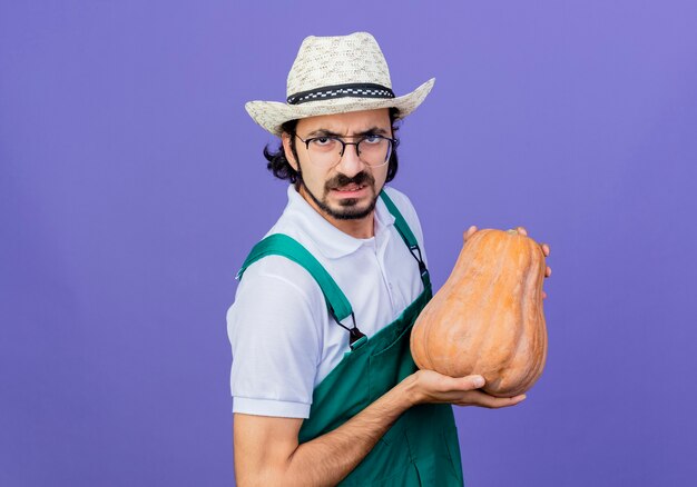 Jeune homme de jardinier barbu portant combinaison et chapeau tenant la citrouille à l'avant avec le visage en colère debout sur le mur bleu