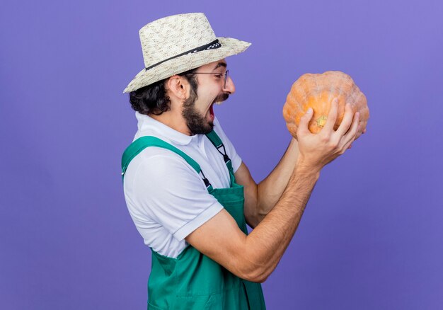 Jeune homme de jardinier barbu portant combinaison et chapeau tenant la citrouille en le regardant heureux et excité debout sur le mur bleu