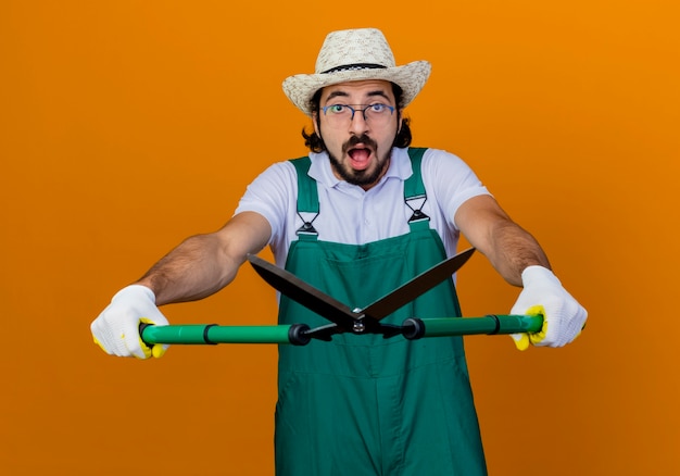 Jeune homme de jardinier barbu portant une combinaison et un chapeau tenant un coupe-haie à l'avant d'être confus et surpris debout sur un mur orange