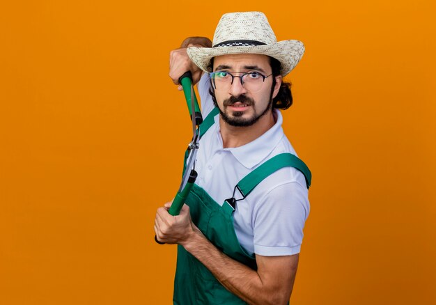 Jeune homme de jardinier barbu portant une combinaison et un chapeau tenant un coupe-haie à l'avant inquiet debout sur un mur orange