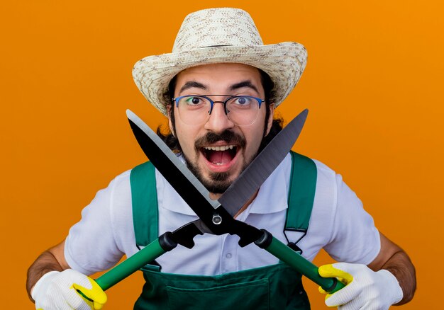 Jeune homme de jardinier barbu portant une combinaison et un chapeau tenant un coupe-haie à l'avant souriant avec un visage heureux debout sur un mur orange