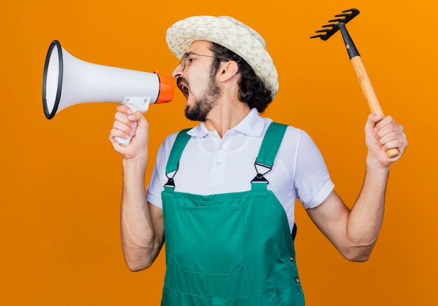 Jeune homme de jardinier barbu portant une combinaison et un chapeau tenant un mini râteau criant au mégaphone en colère debout sur un mur orange