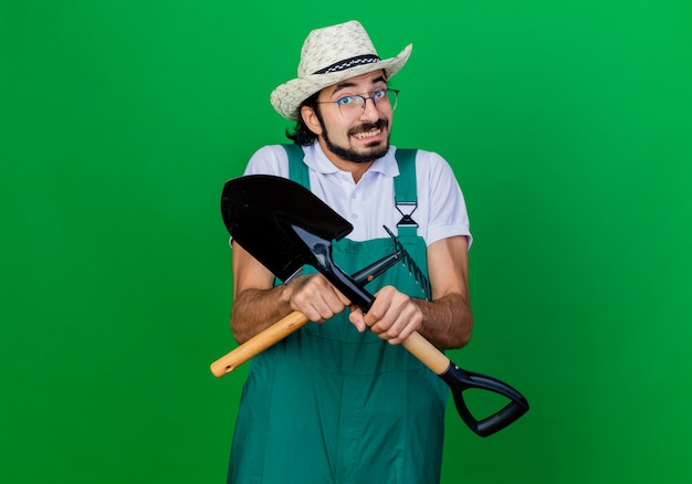 Jeune homme de jardinier barbu portant une combinaison et un chapeau tenant un mini râteau et une pelle confus