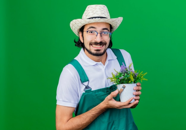 Jeune homme jardinier barbu portant une combinaison et un chapeau tenant une plante en pot à la recherche de sourire avec un visage heureux debout sur fond vert