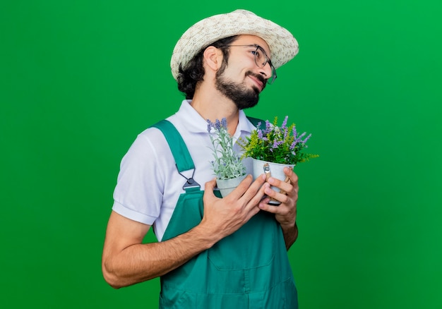 Jeune homme de jardinier barbu portant combinaison et chapeau tenant des plantes en pot ressentant des émotions positives