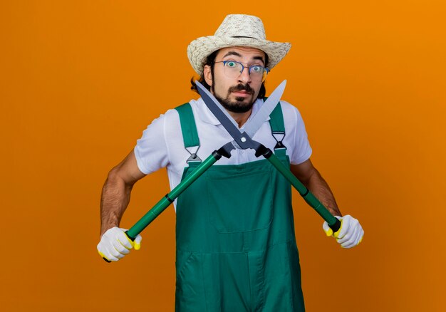 Jeune homme de jardinier barbu portant combinaison et chapeau tenant une tondeuse à haies lookign confus