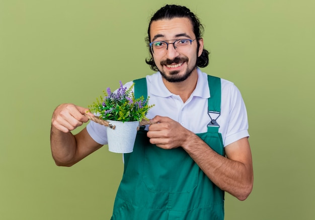 Jeune homme de jardinier barbu portant combinaison montrant plante en pot souriant à l'avant debout sur un mur vert clair