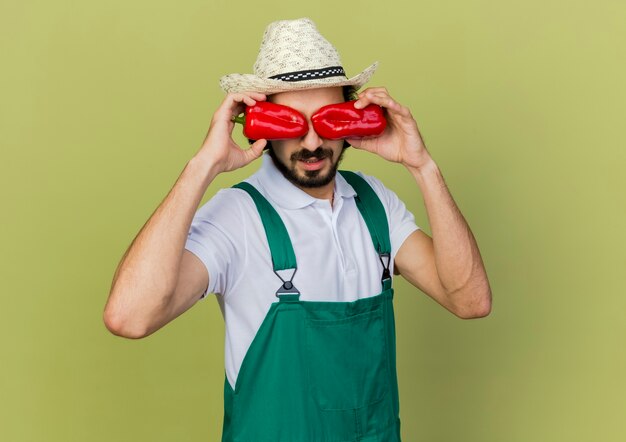 Jeune homme jardinier dans des lunettes optiques portant chapeau de jardinage ferme les yeux avec des poivrons rouges