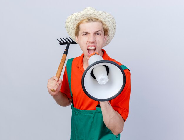 Jeune homme jardinier portant une combinaison et un chapeau balançant un mini râteau criant au mégaphone avec une expression de colère