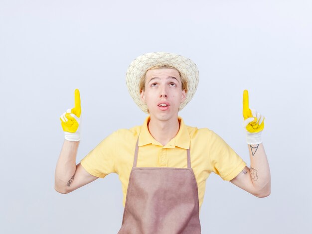 Jeune homme jardinier portant une combinaison et un chapeau dans des gants en caoutchouc pointant avec l'index vers le haut