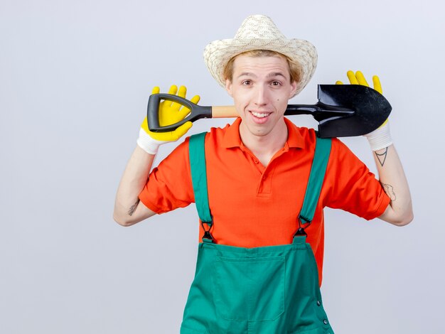 Jeune homme de jardinier portant une combinaison et un chapeau dans des gants en caoutchouc tenant une pelle regardant la caméra en souriant joyeusement debout sur fond blanc