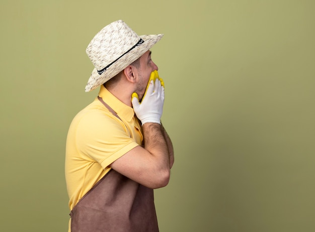 Jeune homme de jardinier portant combinaison et chapeau dans des gants de travail à côté d'être choqué couvrant la bouche avec les mains