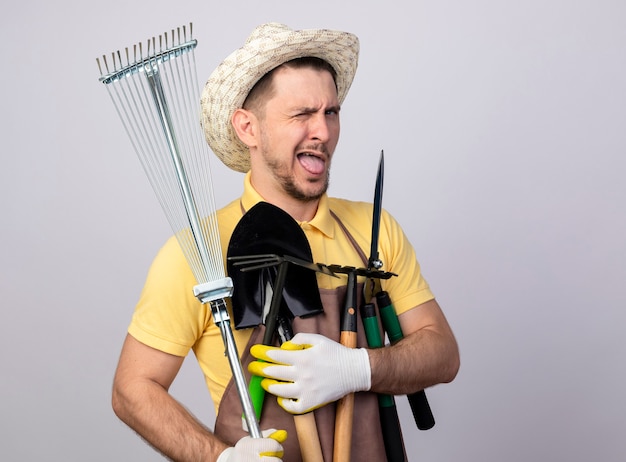 Photo gratuite jeune homme de jardinier portant combinaison et chapeau dans des gants de travail tenant des équipements de jardinage souriant et clignant de la langue