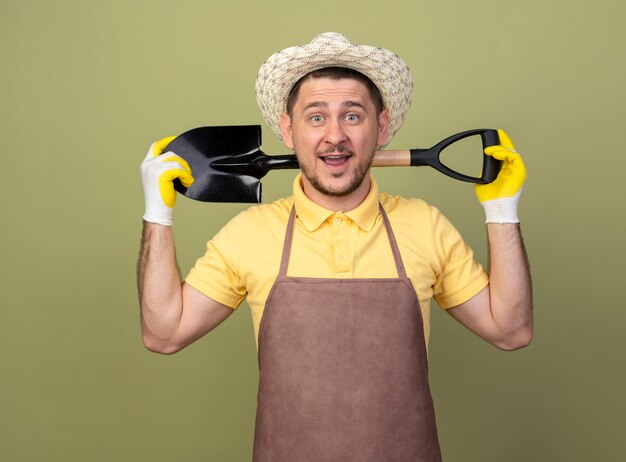 Jeune homme de jardinier portant une combinaison et un chapeau dans des gants de travail tenant une pelle souriant avec un visage heureux