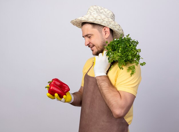 Jeune homme de jardinier portant combinaison et chapeau en gants de travail tenant des poivrons rouges et des herbes fraîches à la recherche avec le sourire sur le visage