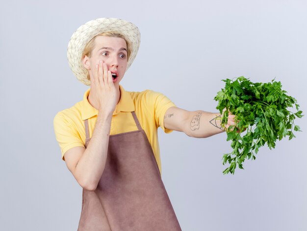 Jeune homme jardinier portant une combinaison et un chapeau montrant des herbes fraîches à côté surpris et étonné