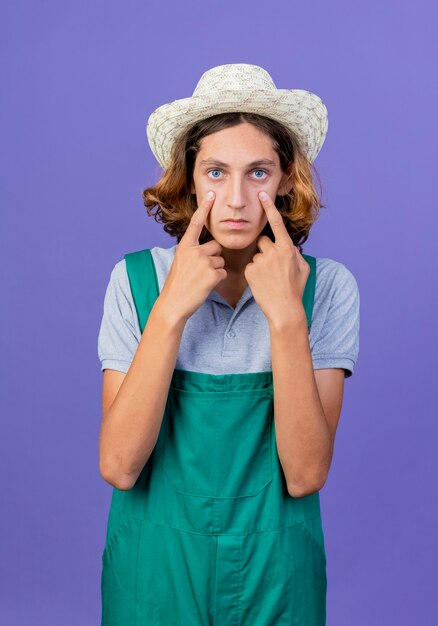 Jeune homme de jardinier portant combinaison et chapeau pointant avec l'index