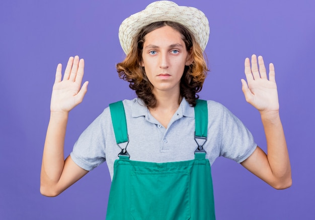 Photo gratuite jeune homme de jardinier portant combinaison et chapeau soulevant des paumes avec un visage sérieux