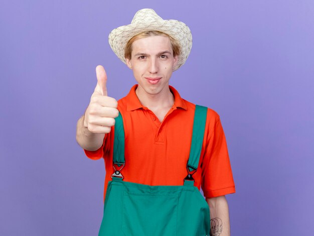 Jeune homme de jardinier portant combinaison et chapeau souriant confiant montrant les pouces vers le haut