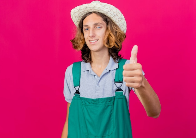 Jeune homme de jardinier portant combinaison et chapeau souriant montrant les pouces vers le haut