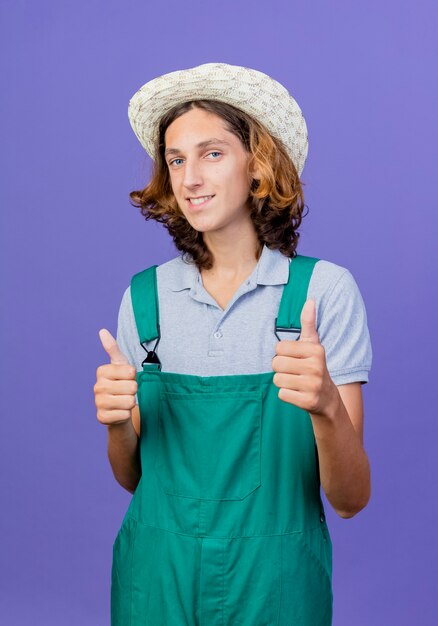 Jeune homme de jardinier portant une combinaison et un chapeau souriant montrant les pouces vers le haut